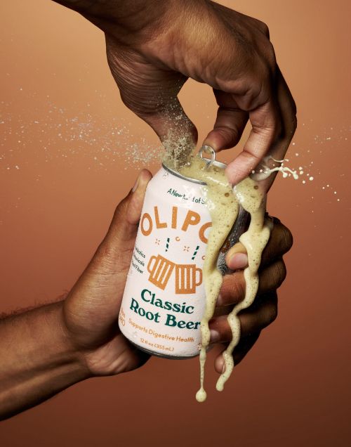 Hands opening a can of Olipop Classic Root Beer, showing the bubbling liquid and splashing against a tan background.