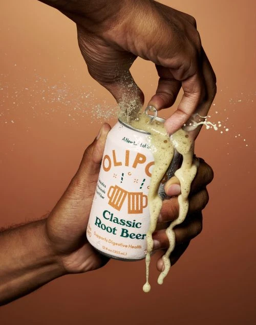 Hands opening a can of Olipop Classic Root Beer, showing the bubbling liquid and splashing against a tan background.