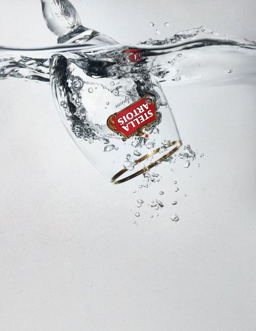 Stella Artois beer glass submerged in clear water with bubbles, gold rim, and red logo against a light gray background.