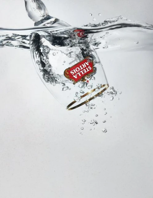 Stella Artois beer glass submerged in clear water with bubbles, gold rim, and red logo against a light gray background.