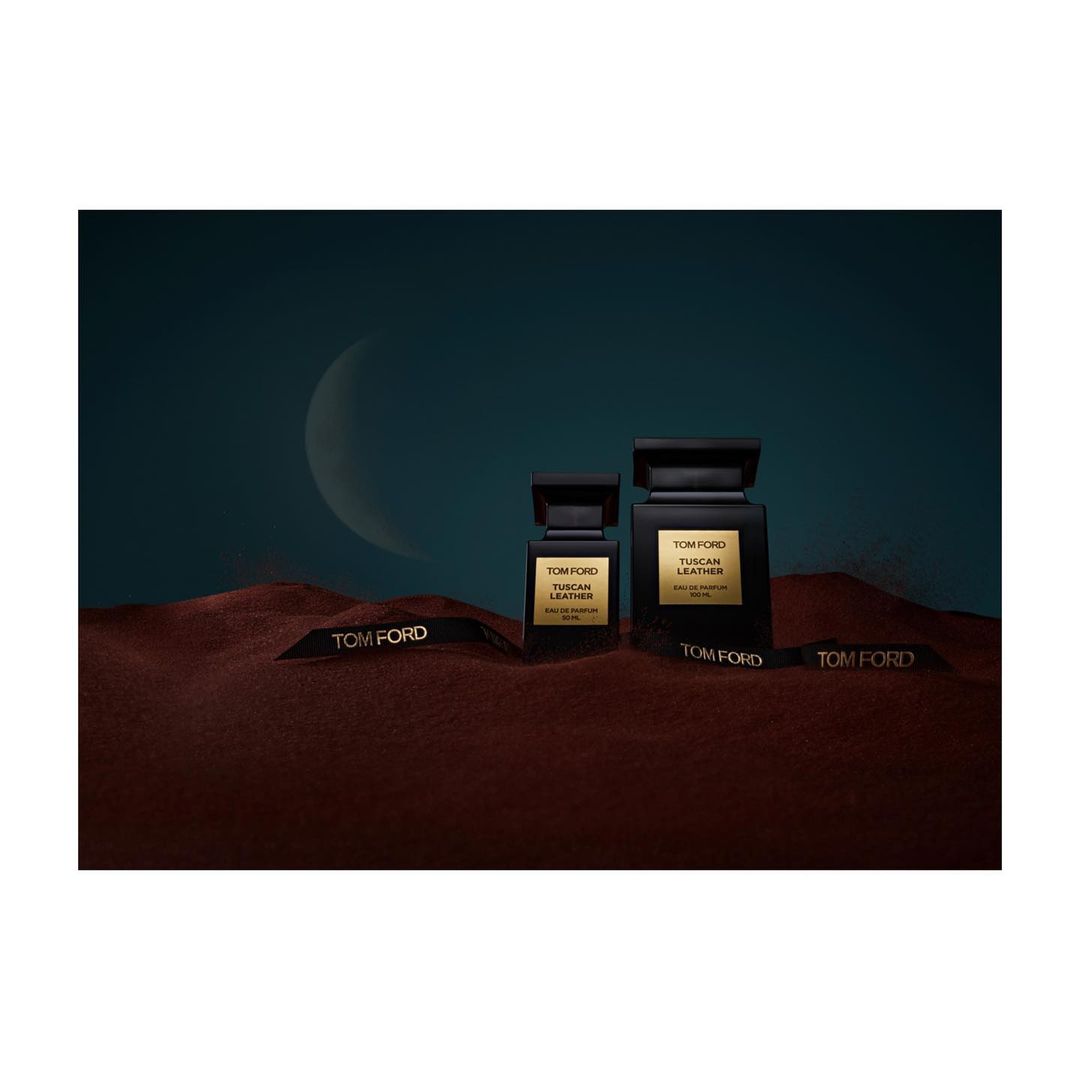 Two Tom Ford Tuscan Leather perfume bottles sit on a textured surface with branded ribbons, set against a teal and crescent moon backdrop.