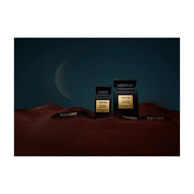 Two Tom Ford Tuscan Leather perfume bottles sit on a textured surface with branded ribbons, set against a teal and crescent moon backdrop.