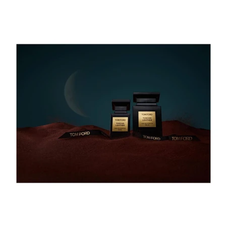 Two Tom Ford Tuscan Leather perfume bottles sit on a textured surface with branded ribbons, set against a teal and crescent moon backdrop.
