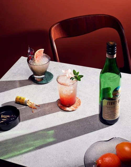 Two cocktails with garnishes and a bottle of liquor on a table with an ashtray, matches, and oranges against a terracotta wall.