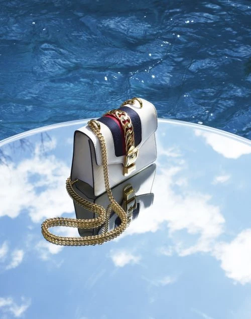 White leather Gucci handbag with a gold chain strap on a reflective round table, background showing water reflections.
