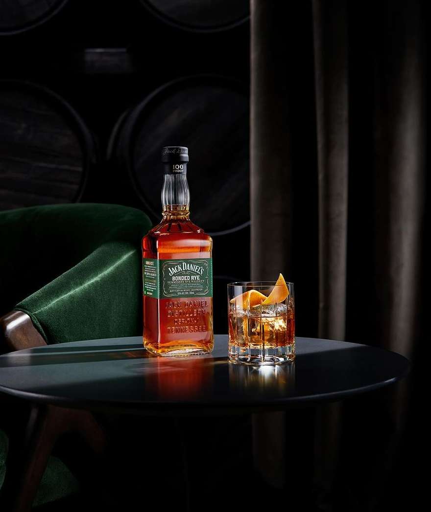 Bottle of Jack Daniel's Bonded Rye whiskey alongside a glass of whiskey with an orange peel on a table in a dark setting.