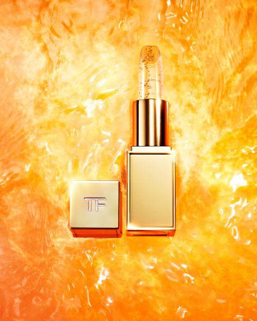Close-up of Tom Ford lipstick with gold flecks. The gold lipstick tube with the cap off, is set against a blurred orange background.