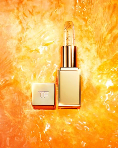 Close-up of Tom Ford lipstick with gold flecks. The gold lipstick tube with the cap off, is set against a blurred orange background.