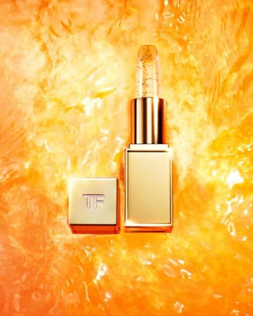 Close-up of Tom Ford lipstick with gold flecks. The gold lipstick tube with the cap off, is set against a blurred orange background.