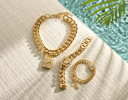 Gold chain necklace with padlock charm, watch, and ring on textured fabric near pool with palm fronds, showcasing luxury jewelry.