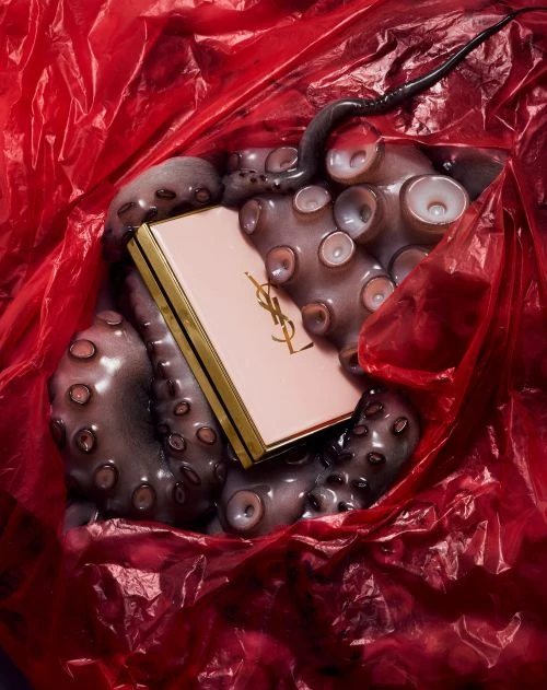 Close-up of makeup compact on octopus tentacles in red plastic bag, shot with studio lighting for fashion and beauty context.
