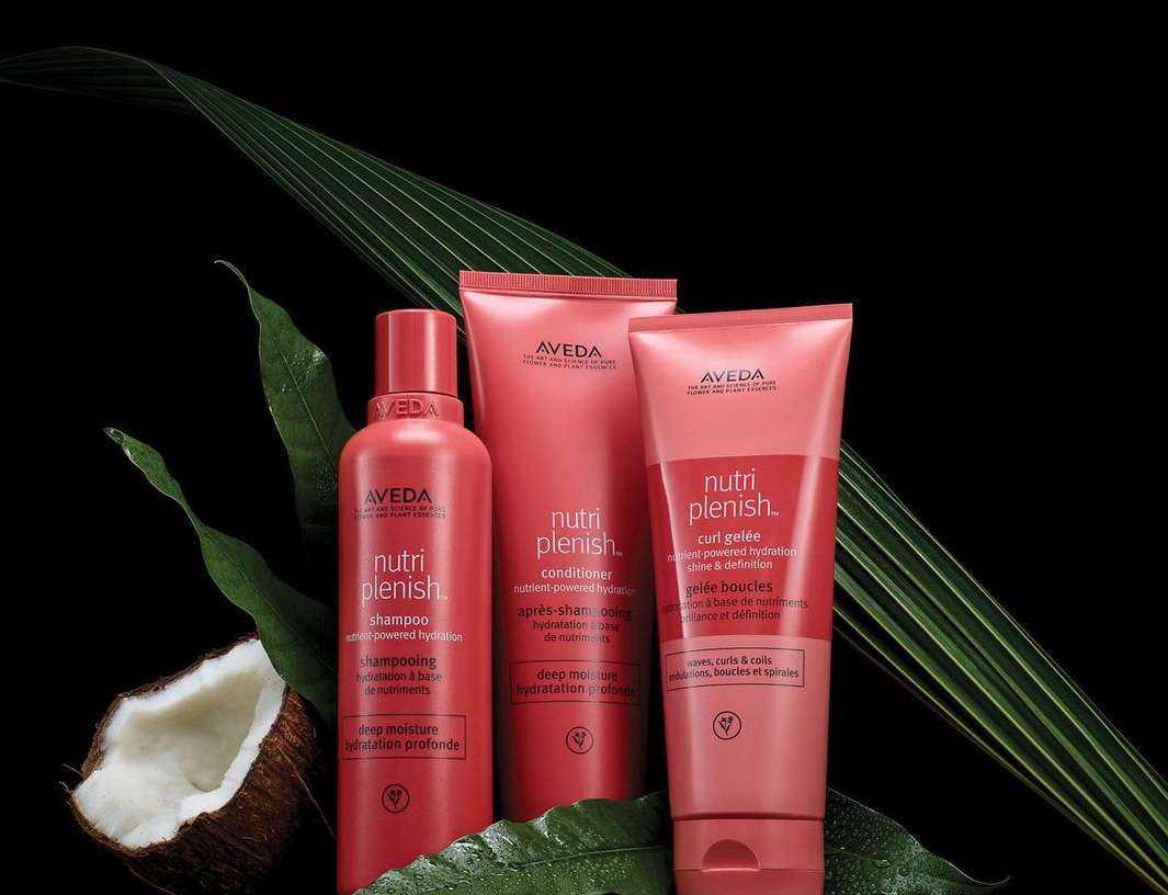 Aveda Nutriplenish hair products including shampoo, conditioner, and curl gelée, staged with coconut and tropical leaf elements against a black background.
