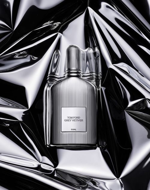 Tom Ford Grey Vetiver fragrance bottle with a ribbed design, front-center on a reflective, crumpled silver surface, studio lighting.