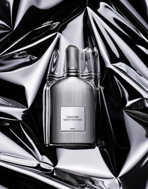 Tom Ford Grey Vetiver fragrance bottle with a ribbed design, front-center on a reflective, crumpled silver surface, studio lighting.