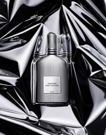 Tom Ford Grey Vetiver fragrance bottle with a ribbed design, front-center on a reflective, crumpled silver surface, studio lighting.