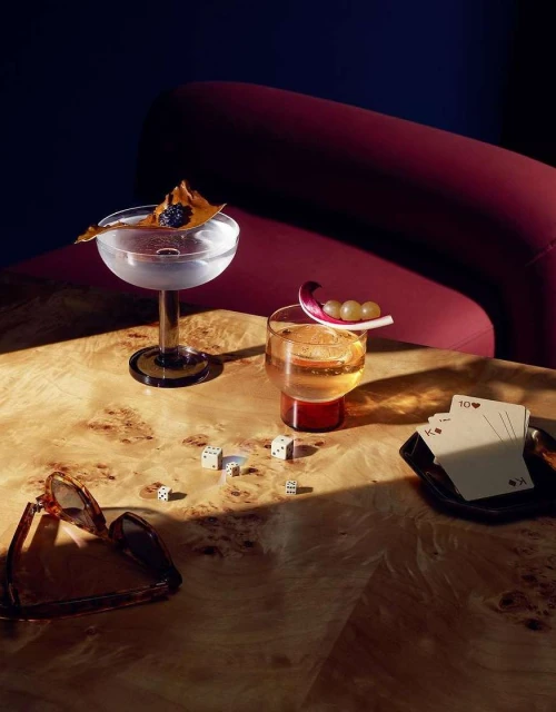 Two cocktails garnished with fruit and a leaf on a wood table with playing cards, dice, and sunglasses in a dimly lit setting.