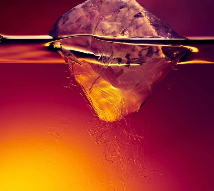 A close-up shot captures an ice cube melting in a glass of liquid, creating swirling patterns and a gradient of amber and red hues.