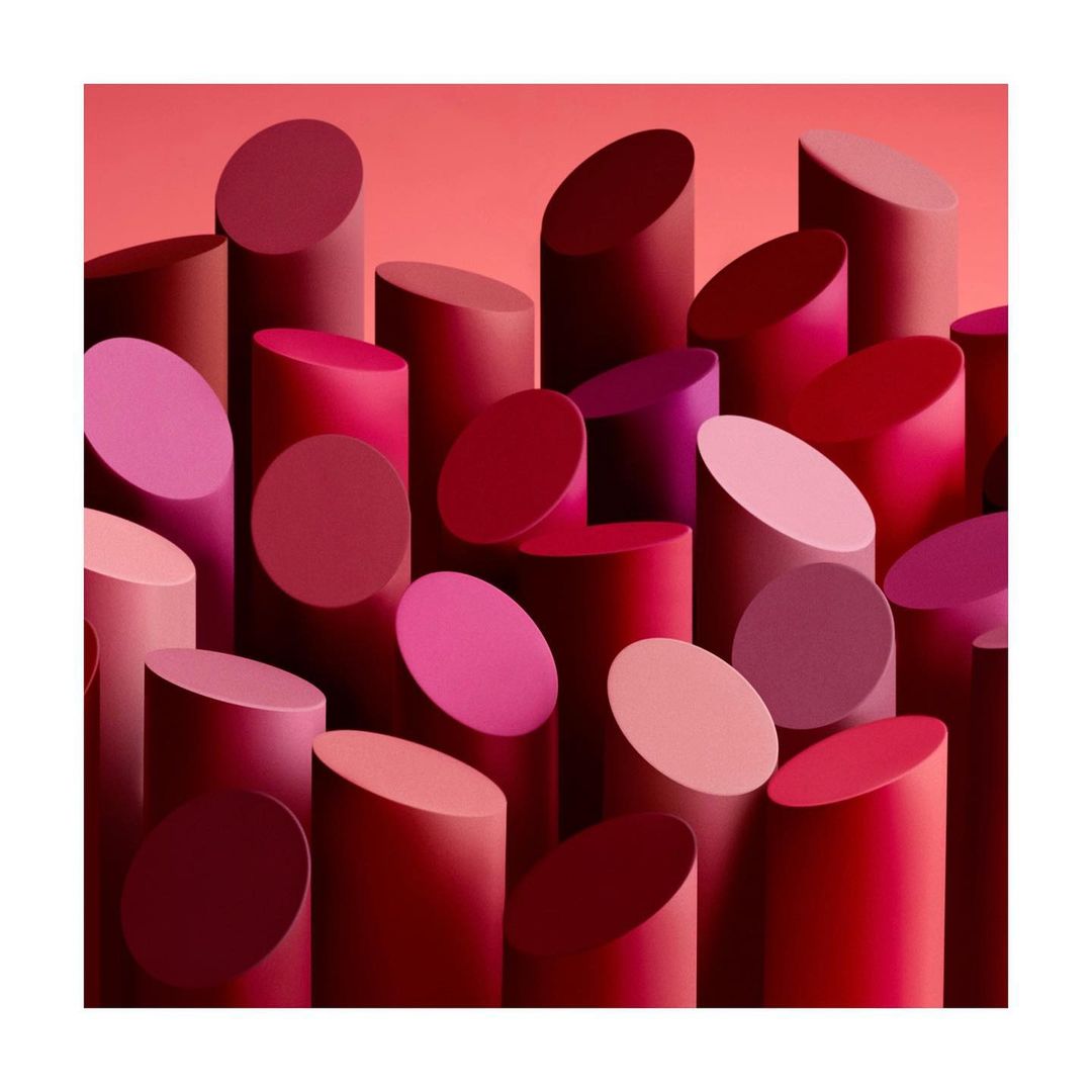 Close-up arrangement of multiple cylindrical lipstick tubes in varying shades of red, pink, and burgundy against a pink background.