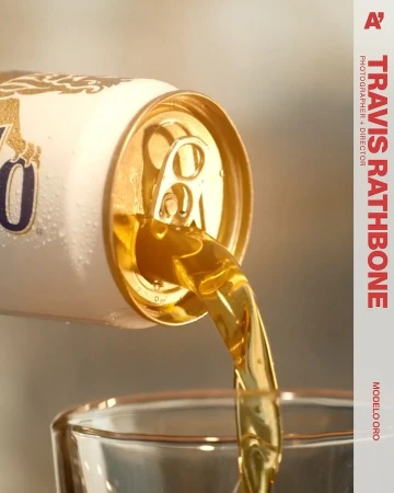 Close-up of Modelo Oro beer pouring from a white and gold can into a glass, highlighting the beverage's golden hue and refreshing texture.
