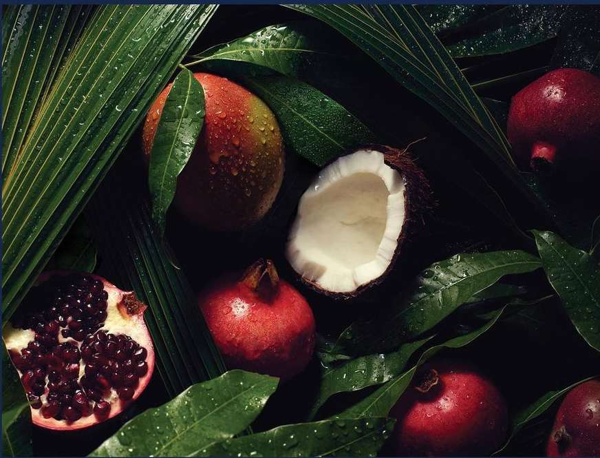 Overhead shot of pomegranates, a coconut, and a mango with water droplets on lush green tropical leaves creates a vibrant, fresh still life.