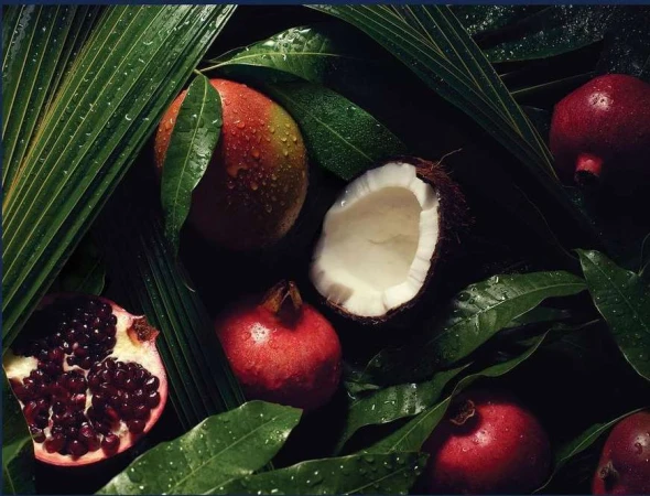 Overhead shot of pomegranates, a coconut, and a mango with water droplets on lush green tropical leaves creates a vibrant, fresh still life.
