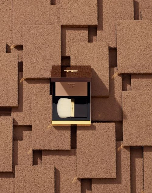 Tom Ford beauty product bronzer compact with a brush, presented against a textured tan geometric background.