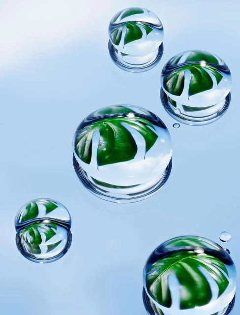 Macro photography showcasing water droplets with reflections of green leaves on a light blue background, creating a visually striking nature image.