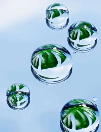 Macro photography showcasing water droplets with reflections of green leaves on a light blue background, creating a visually striking nature image.