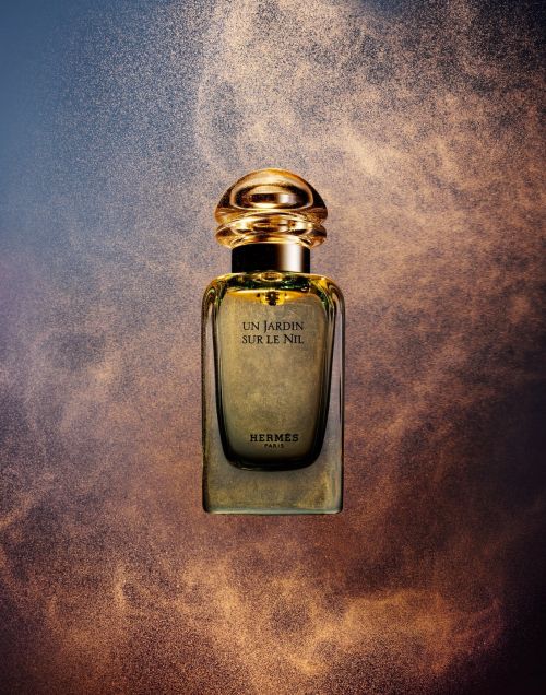 Elegant Hermès perfume bottle, Un Jardin Sur Le Nil, stands centered amidst a golden glitter dust cloud on a blue-beige backdrop.