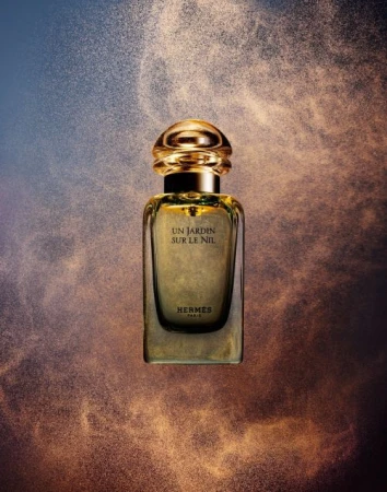 Elegant Hermès perfume bottle, Un Jardin Sur Le Nil, stands centered amidst a golden glitter dust cloud on a blue-beige backdrop.