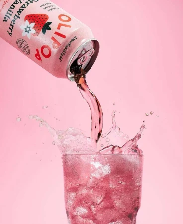 Olipop Strawberry Vanilla drink being poured into a glass of ice against a pink background. Refreshing, fizzy, and visually appealing.