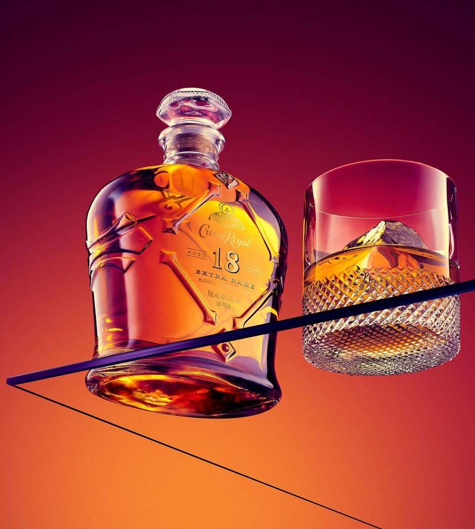 A bottle of Crown Royal 18 and a glass of whiskey with an ice cube are displayed against a red gradient background in a studio.