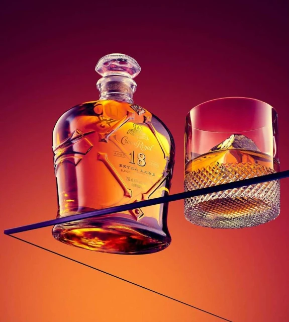 A bottle of Crown Royal 18 and a glass of whiskey with an ice cube are displayed against a red gradient background in a studio.