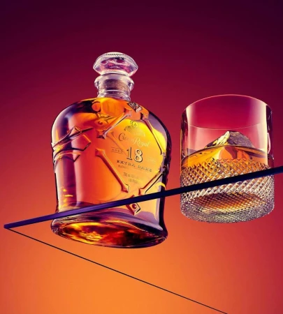 A bottle of Crown Royal 18 and a glass of whiskey with an ice cube are displayed against a red gradient background in a studio.