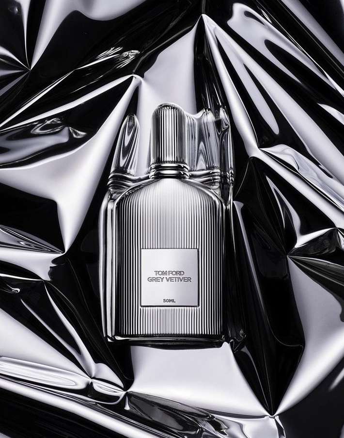 Close-up shot of a Tom Ford Grey Vetiver perfume bottle against a reflective, crumpled silver background, studio lighting.