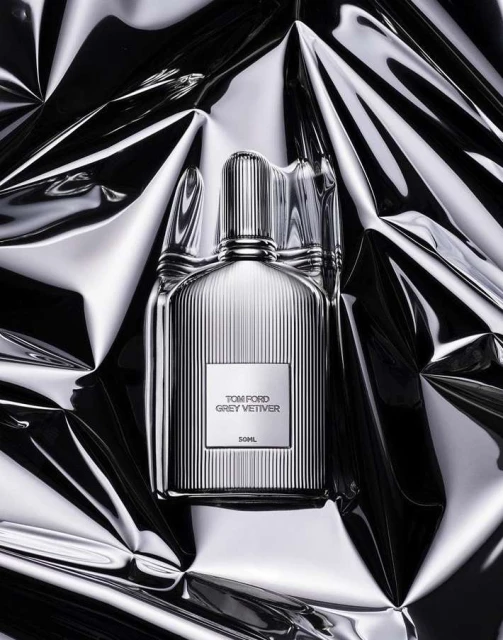Close-up shot of a Tom Ford Grey Vetiver perfume bottle against a reflective, crumpled silver background, studio lighting.