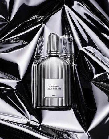 Close-up shot of a Tom Ford Grey Vetiver perfume bottle against a reflective, crumpled silver background, studio lighting.