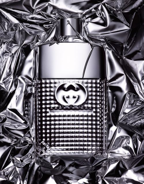 Close-up studio shot of a Gucci Guilty cologne bottle. The silver bottle is surrounded by crumpled aluminum foil for a textured background.