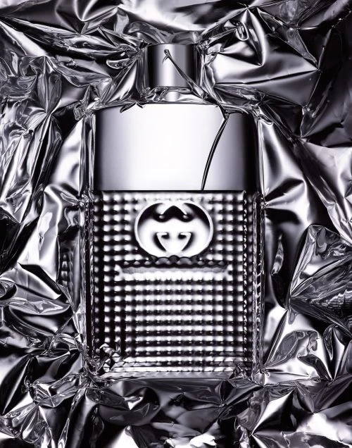 Close-up studio shot of a Gucci Guilty cologne bottle. The silver bottle is surrounded by crumpled aluminum foil for a textured background.