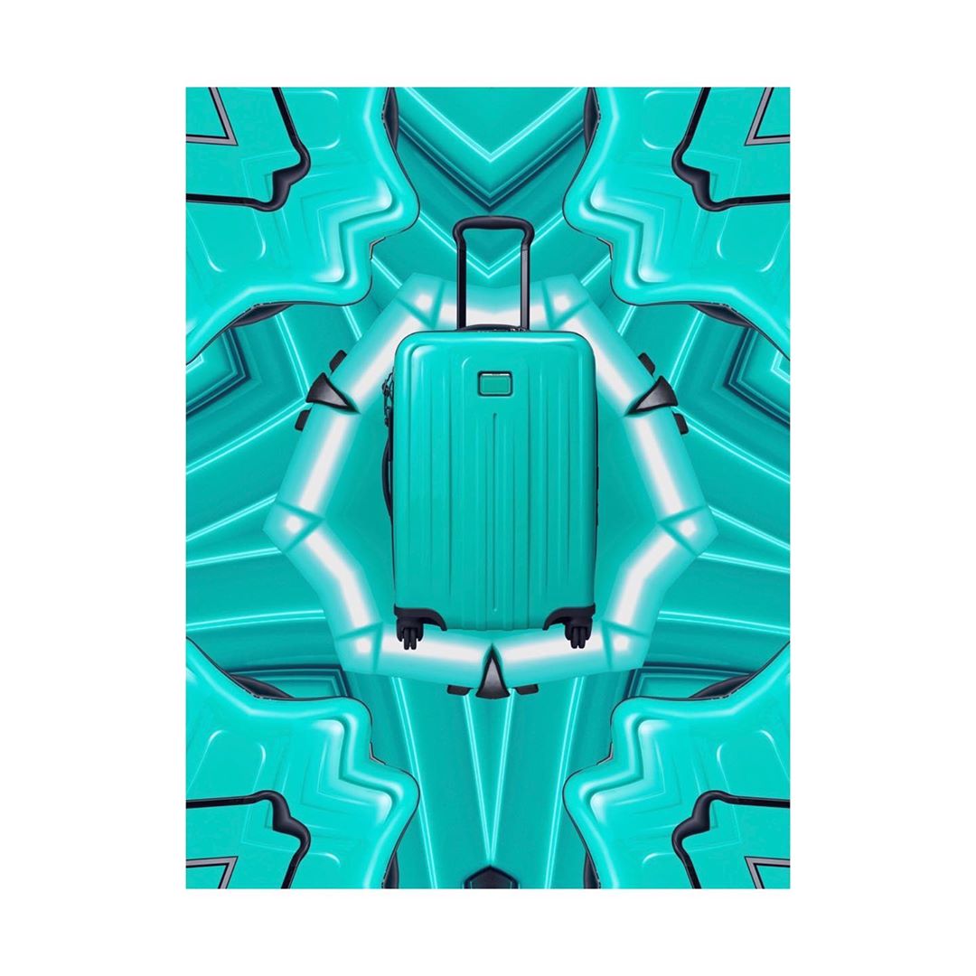 Studio shot of a turquoise hard-shell suitcase with a telescopic handle, set against an abstract turquoise and white patterned backdrop.