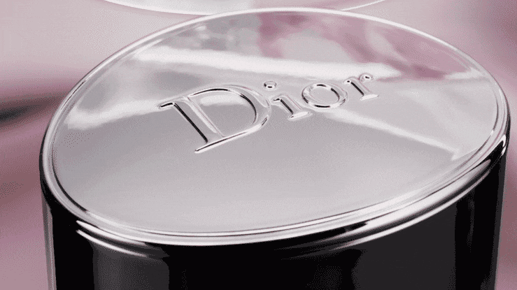 Close-up of a glossy metallic cosmetic cap with the embossed Dior logo against a soft pink background.