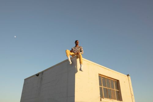 An adult male is sitting on the roof of a white building against a clear blue sky. He is wearing a patterned shirt and light-colored pants.
