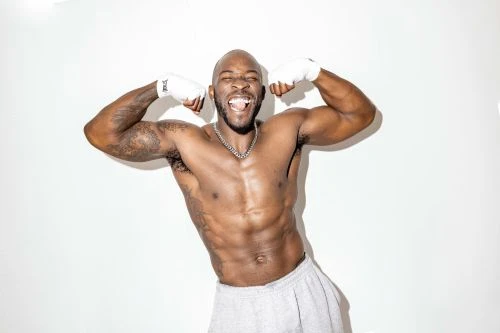 Shirtless African American man with arm wraps flexes biceps against a white backdrop, showing muscular build in studio lighting.