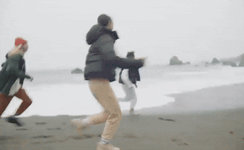 Four young adults running on a beach. They are wearing jackets and pants. The beach has dark sand and the ocean waves are visible.
