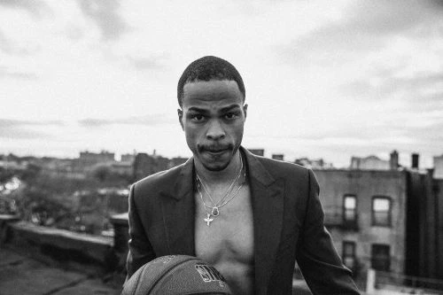 Black man holding a basketball wearing an unbuttoned blazer and layered necklaces on a rooftop in Los Angeles.
