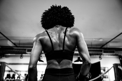 Female boxer with muscular back standing in a boxing ring, wearing black sports bra and boxing gloves in black and white photography.