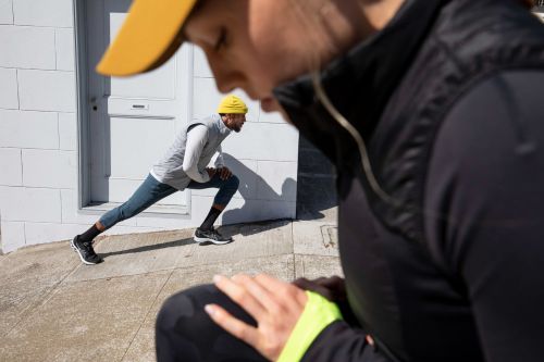 Two men stretch outdoors on concrete; one in black in foreground, another stretches by the building in hat and vest.