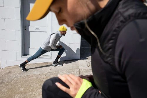 Two men stretch outdoors on concrete; one in black in foreground, another stretches by the building in hat and vest.