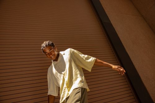 Young adult male in casual shirt and olive pants smiles, posing in front of a textured brown building exterior on a sunny day.