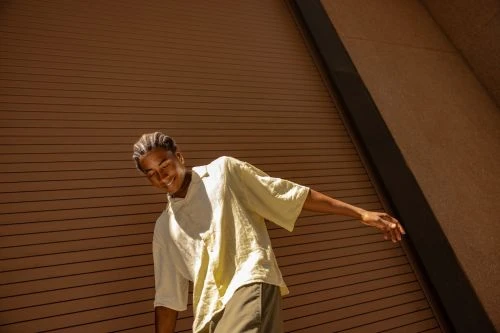 Young adult male in casual shirt and olive pants smiles, posing in front of a textured brown building exterior on a sunny day.
